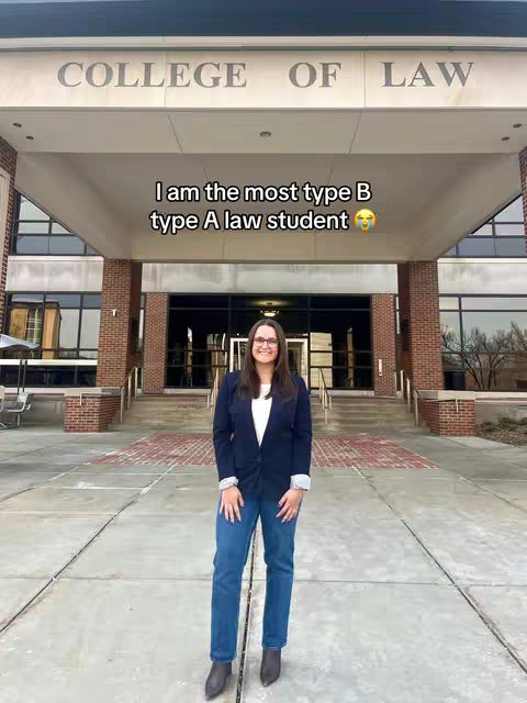A woman in jeans and a blazer stands in front of a college building. Text overlayed on the image reads, "I am the most type B type A law student".