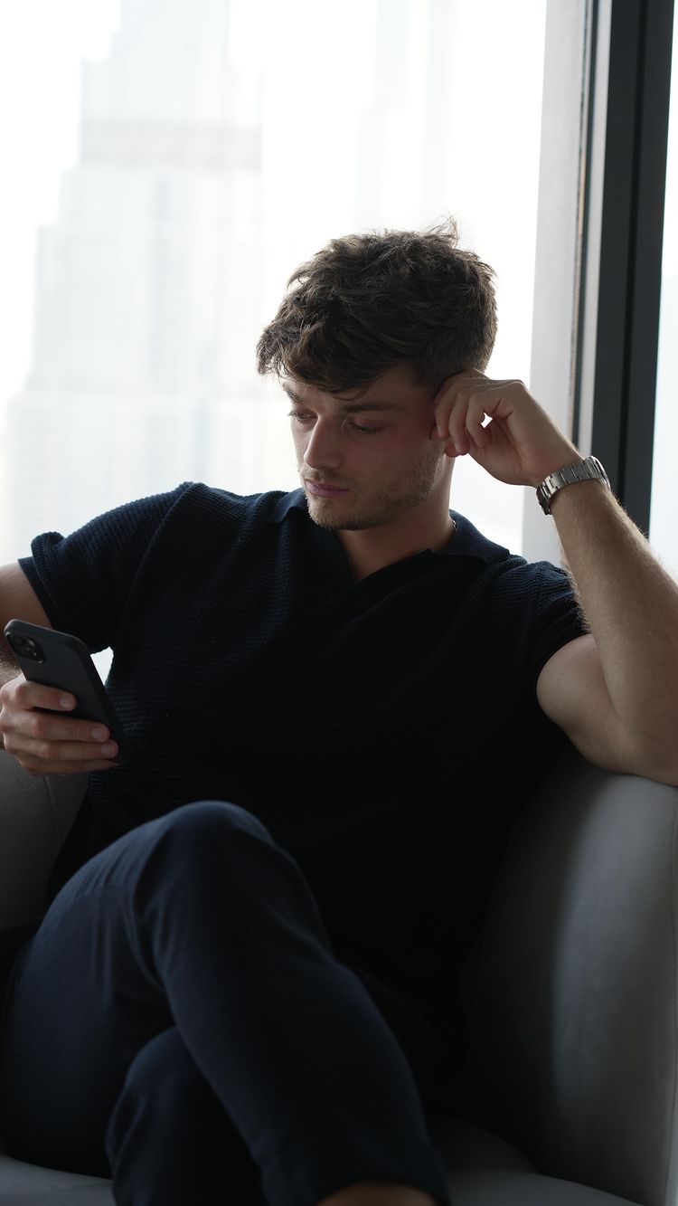 A man in a navy polo shirt and dark pants sits in a modern chair, looking at his phone. The background includes a large window with a view of high-rise buildings. He rests one hand on his temple. The lighting is soft and natural.