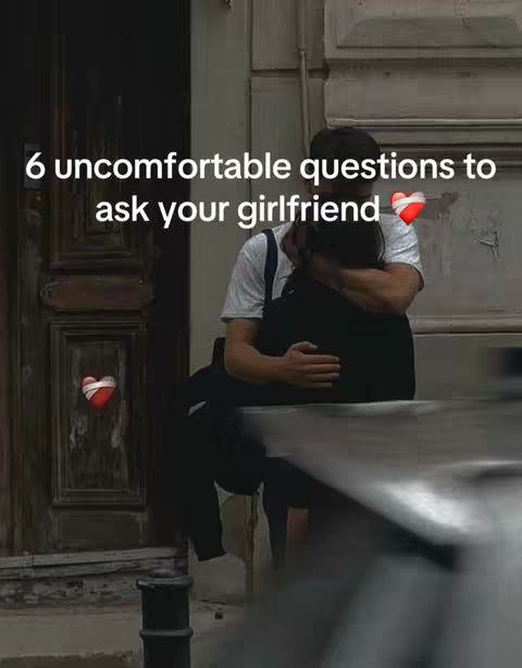 A couple is embracing in a street scene. Text overlay indicates "6 uncomfortable questions to ask your girlfriend". The scene features a man in a white t-shirt embracing a person with dark hair. A heart is placed near the overlay text.