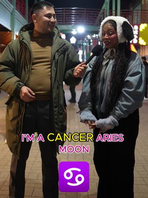 An outdoor scene at night, with two people facing each other, talking. One figure is wearing an olive green jacket and the other is wearing a white hat. A text overlay at the bottom has astrological signs.
