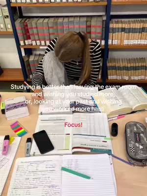 The image shows a person at a desk, likely studying with an open book and notes. The background has bookshelves filled with books. The person is seen from above, with their head down and a part of their back visible. A text overlay reads "FOCUS!"