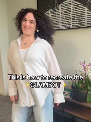 A woman with curly hair stands indoors, smiling at the camera. She's wearing a white shirt over a white undershirt and light blue jeans. Text overlayed on the image says 'This is how to recreate the GLAMBOT'. A stylish painting hangs on the wall behind her, and a table with plants and decoration is on the right.
