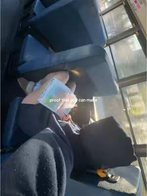 The image shows a person lying down on a bus seat by a window. The person is covered with a hoodie and a blanket, and is holding a notebook or planner. The seats are blue, with a yellow object in view.