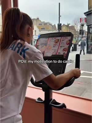 A person is shown from the back, exercising on a stationary bike while looking at a screen displaying a KFC menu. The image is taken in a gym with a street visible through the window. The KFC logo can be seen in the background.
