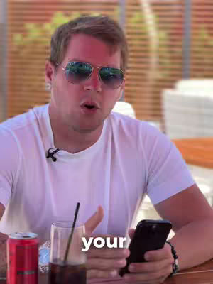 A man wearing sunglasses is holding a phone and speaking. He is at a venue with a blurred background with a beverage nearby. There is a hook that states 'your'.