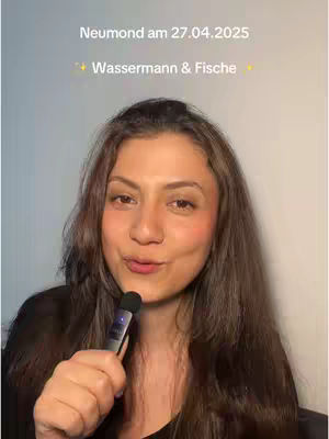 A woman with long brown hair is holding a microphone and looking at the camera. She has light makeup on and there is text overlayed in white on the upper part of the image.