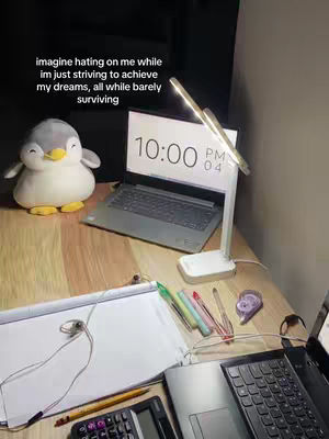 The image shows a desktop workspace. A laptop with a lit screen displaying '10:00 PM 04' sits on a wooden surface, next to the laptop is a white desk lamp. To the left, a plush penguin toy faces the laptop. Below the laptop are school/work supplies like pens, a sketchbook, a calculator, and headphones. The background is dark.