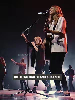 The image captures a live performance with two women on stage singing and playing instruments. One person plays the guitar and sings, while the other sings into a microphone, both engaged in worship, with a stage and a background of other people.