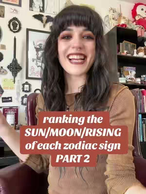 A woman with dark hair and bangs smiles at the camera. She is wearing a brown top. A text box overlays the image that says, "ranking the SUN/MOON/RISING of each zodiac sign PART 2". The background includes a wall with pictures, shelves, and a small red doll.
