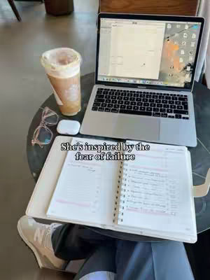 A top-down view captures a workspace: a laptop displaying a document, a notebook with handwritten notes, a drink in a paper cup, and glasses. The setting appears to be a cafe or similar environment captured in natural light.