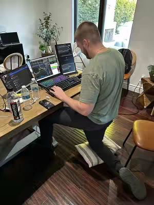 A man is working from home, sitting at a desk with multiple computer screens. He is dressed in a green t-shirt and black pants. He appears to be working from his knees up on an office style desk, angled towards the camera. Various items cover the desk, including two computers.