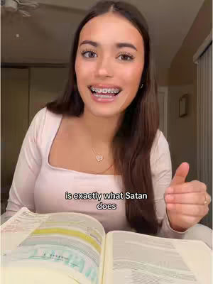 A young woman with long brown hair and braces is smiling and giving a thumbs-up. She is wearing a light pink top and a necklace with a heart pendant. She is sitting in front of an open book, likely a Bible, and there's text overlayed on the image.