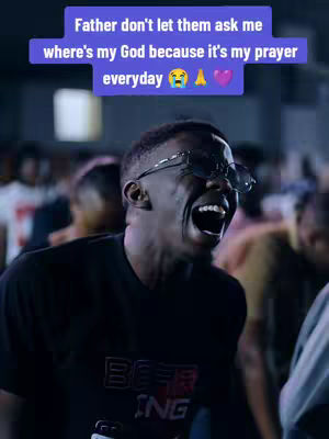 A medium shot of a Black man with short hair, wearing a black t-shirt and sunglasses. He is mid-scream, mouth wide open, showcasing white teeth. The background is a blur of people, suggesting a crowd or audience. A purple text overlay appears above him.