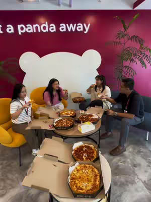 A group of four people are seated around a round table, sharing a meal that prominently features pizzas. The setting is indoors, in a modern space with a red wall adorned with a white bear logo, suggesting a commercial location. The lighting appears to be bright, and everyone seems to be enjoying themselves. Pizzas in cardboard boxes are visible, along with drinks, suggesting a casual meal atmosphere.