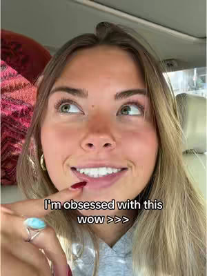 A young woman is looking up and to the side with a happy expression. The overlay text reads 'I'm obsessed with this wow >>>'. She is inside of a car. She is wearing a ring with a teal gem, and has painted nails.