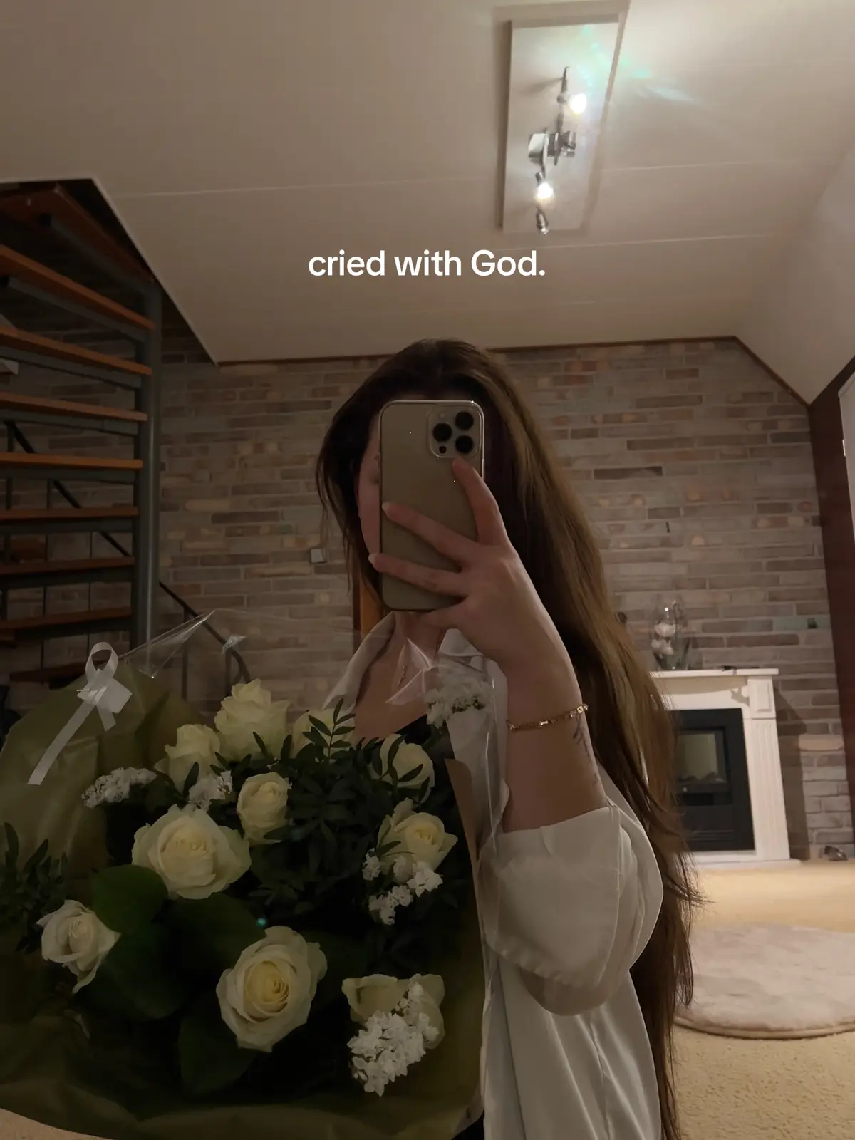 A person is taking a selfie while holding a bouquet of white roses. Her face is partially obscured by the phone, but her long brown hair is visible. She is wearing a white shirt and a gold bracelet. The background shows a living room with a brick wall, a fireplace, and a spiral staircase.