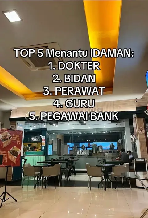 The cover image shows a brightly lit interior space, possibly a cafe or restaurant, with a list of "TOP 5 Menantu IDAMAN" (Ideal Son-in-law) in Indonesian displayed as text overlay. The list features Doctor, Midwife, Nurse, Teacher, and Bank Employee. The composition is mid-shot, capturing the interior setup with tables, chairs, interior decor, and the text in the center makes up the image's focus.