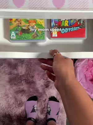 The image shows the lower half of a person's body with legs and feet. The person is wearing light purple socks with a black and white design. They are standing on a fluffy, light purple rug. In the background, there's a shelf with two video game cases. Above, a shelf has pink shapes with hearts.