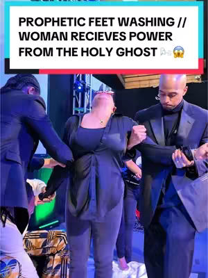 The image shows a woman supported by two men as what appears to be some sort of religious ritual. The woman looks up with her arms raised. The men are dressed in suits and appear to be of African descent. The background consists of an indoor stage setup lit with blue and green lights.