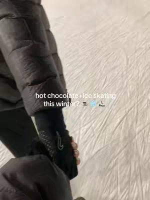 A close-up shot of someone's gloved hand holding onto another person's arm or possibly a jacket sleeve. The background is a white, textured surface, presumably ice. Text overlay reads "hot chocolate + ice skating this winter?" followed by some emojis.