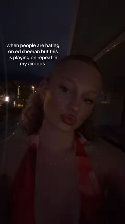 The image is a selfie of a young woman taken in a dark environment, possibly a car or indoor space at night. She is facing the camera with a slight pout, and her lips are emphasized with red lipstick. Her hair is styled, and she appears to be wearing a red top. The background is blurred, suggesting movement or low lighting. The text overlay addresses the topic of Ed Sheeran's music.
