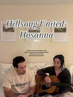 The image features a couple with musical instruments. The woman in the foreground is holding a guitar and singing, and the man next to her looks at her while sitting on the bed. The background features framed pictures hanging on the wall.