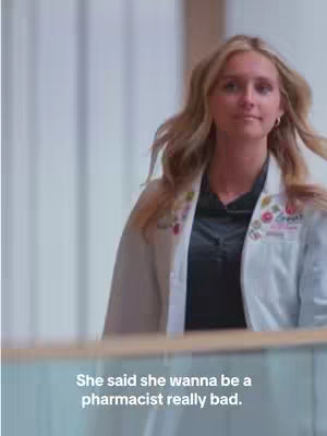 A woman in a white lab coat walks forward in a bright, clean environment. The image captures her from the waist up.