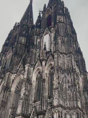 An upward view of the Cologne Cathedral shows its towering, dark gray facade against a pale, overcast sky. The architectural details are intricate, with pointed arches, tall spires, and numerous ornate elements creating a sense of grand scale and historical significance.