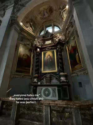 The image shows the interior of a church. The composition is centered on a religious shrine, with an ornate structure topped by a picture of a saint. On either side, there are paintings, and the high arched ceiling features painted details. The color palette is dominated by muted earth tones, giving the space a solemn and historic feeling.