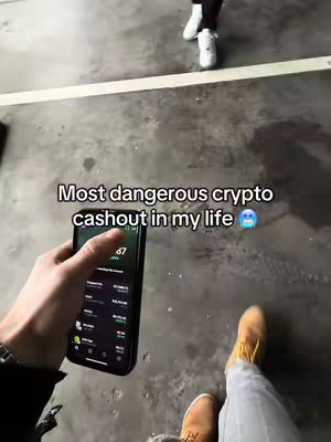 The image shows a first-person perspective of someone holding a smartphone displaying a crypto app. The person is pointing at the screen. The foreground shows the person's hand, a watch, and part of their clothing. In the blurred background, another person's legs and the upper part of their body are visible. The scene is set in a gray, possibly concrete, environment.