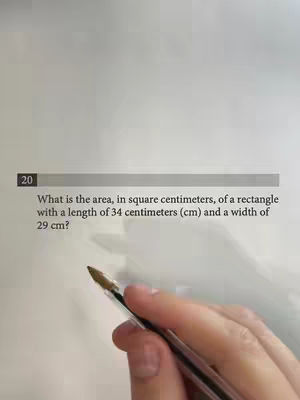 The image shows a sheet of paper with a math problem question about finding the area of a rectangle, displayed in a formal layout. A hand holding a pen gestures toward the question, with focus on the text and calculations. The background is a plain, neutral color.