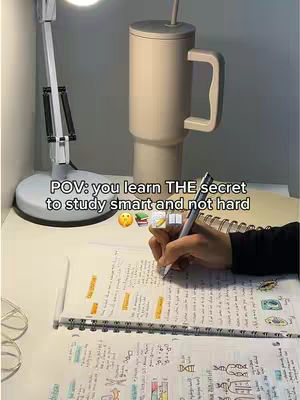 The image is taken from an elevated angle, focusing on a white desk. A notebook with handwritten notes and highlighted text is open and visible, along with a pen resting on the page. To the right, a large, insulated tumbler with a handle sits upright. The background includes a desk lamp and a wall, with the overall scene illuminated by soft lighting. The composition and details suggest a study or work environment.