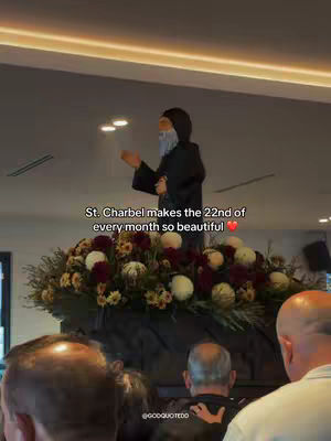 The image shows a statue of St. Charbel in a dark-colored robe with a white beard and a veil, standing atop a platform adorned with a large floral arrangement. The foreground shows the heads of several people, likely an audience. The text overlay reads "St. Charbel makes the 22nd of every month so beautiful" with a heart emoji.