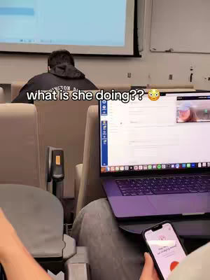 The cover image shows a classroom setting. In the foreground, a person's arm and hand are visible, holding a smartphone. In the middle ground, a laptop is open on a desk, displaying a screen. Behind it, another person appears to be sitting, their back toward the camera, wearing a hooded sweatshirt. In the background, there is a projector screen. The text 'what is she doing??' is overlaid on the image.