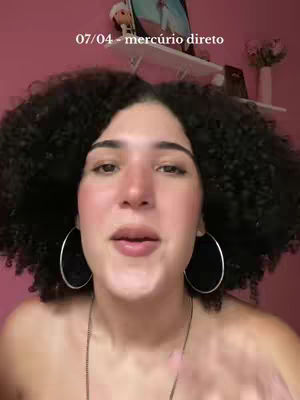 A woman with dark, curly hair framed around her face is centered in the shot. She is wearing large silver hoop earrings and a delicate chain necklace. The background is a soft pink. Text overlays at the top indicate information.