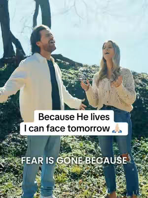 A wide shot captures a man and a woman standing outdoors in a natural setting, likely a park or meadow. The man, on the left, wears a light-colored jacket over a dark shirt and jeans, his expression is joyful as he looks upwards. The woman, on the right, is smiling, wearing a light sweater, jeans and looking upwards, as well. The background features blurred greenery with some tree trunks visible, and the sky is a light blue. There are overlaid text elements, in white, placed near the bottom of the image.