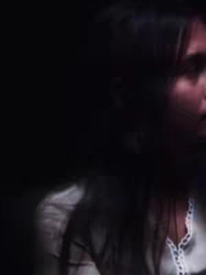 The image shows a person in a close-up shot, with most of the face visible. The background is dark, creating a contrast that makes the person stand out. The lighting appears dim, possibly from a single source, highlighting the person's features and conveying a sense of mystery or intimacy. The person has long dark hair and are wearing a white top with a neckline.