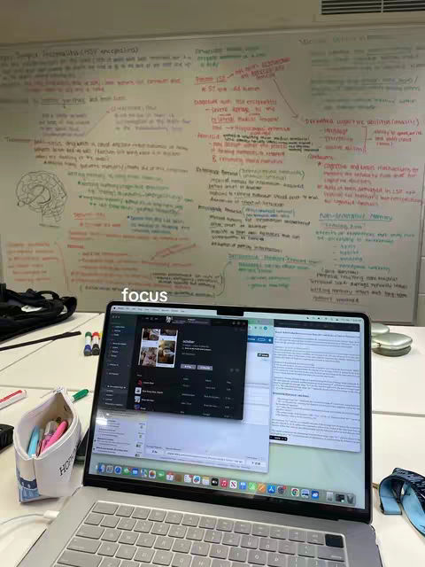 A laptop displaying application screens sits on a desk. In the background, there is a whiteboard with handwritten notes and diagrams, next to a wall. A pencil case and colored markers are visible, suggesting a study environment.