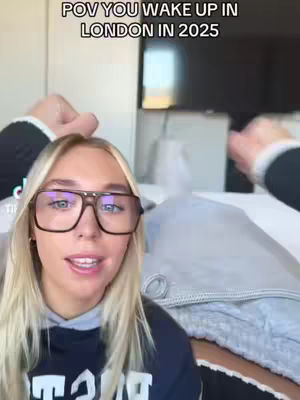 A woman with blonde hair and glasses looks directly at the camera, in a room with a bed and television visible in the background; the text on the cover reads: POV YOU WAKE UP IN LONDON IN 2025.