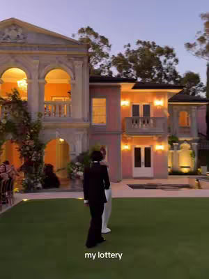An outdoor shot capturing an elaborate architectural scene under an evening sky. The foreground showcases a well-manicured lawn, while the midground presents a multi-level building with ornate designs and warm interior lighting. Two figures, one in a black outfit and the other in white, are walking away from the camera. Text overlayed on the bottom reads 'my lottery'. The color palette features the warm tones of the building lights against the deepening blueness of the sky.