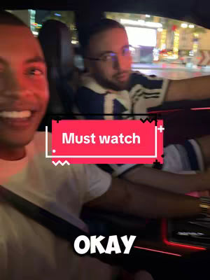 A selfie-style video featuring two men in the front seats of a car at nighttime. The focus is on the man on the left, who is smiling. The background shows city lights and potential buildings, suggesting a cityscape at night. The visual composition is a casual, on-the-go shot, with overlays on the image.