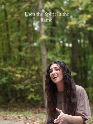 A young woman with long, wavy dark hair sings outdoors. She's wearing a beige t-shirt. The background is a blurry green forest. Text at the top says:'Then the Spirit lit the flame.'