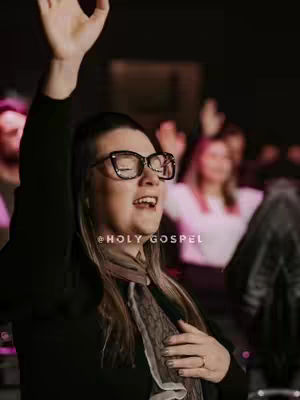 A woman with glasses has her hand raised near her head, presumably worshipping.  She's wearing a dark jacket and a scarf. Other blurred figures are behind her.. The image is set in an indoor space, possibly a church.