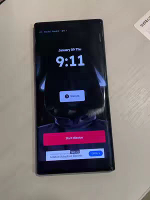 A picture of a black phone which has a dark background, showing a low-angle image of a person wearing a black jacket and a black hood. The phone screen shows the date and time at the top, with the time reading 9:11. There is also a button that says "Start Mission" in red.