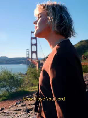 A woman stands on a rocky outcrop, looking towards the Golden Gate Bridge in San Francisco at sunset. The bridge is visible in the background, and the sky is a gradient of orange and blue. The woman is wearing a dark jacket. Overlayed text reads 'How I love You Lord'