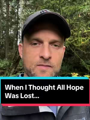 Close-up of a man in his late 30s or early 40s wearing a black baseball cap, looking directly at the camera with a somber expression. The background is blurred greenery, suggesting an outdoor setting. A black bar with white text overlays the bottom of the image.