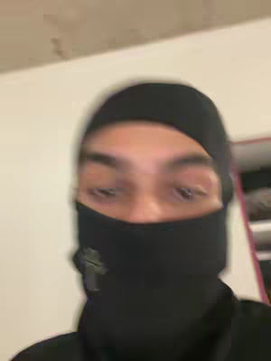 The image is a blurry, close-up shot of a person’s face, focusing on their eyes, partially obscured by a black head covering and face mask. The background is indistinct, with a neutral, possibly indoor setting. The lighting seems to be soft and diffused.
