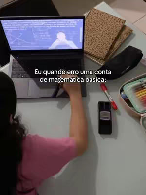 A person is shown from the back, wearing a pink shirt and a black cap, placing her hand on a laptop. The laptop shows a math problem on the screen. Beside the laptop there are notebooks, a case, a red marker, a black phone and some colorful highlighters. The background is a white table.