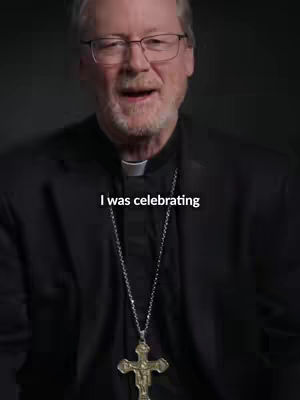 A medium shot features a man with fair skin and glasses. He's wearing a black suit jacket with a white collared shirt and a silver cross necklace. Above the man appears the text overlay. The setting appears to be a dimly lit space with a plain background.