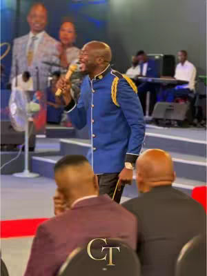 A man in a blue military-style jacket with gold accents is speaking into a microphone on a stage. Other people are visible in the background, which seems to be a church or auditorium setting. The man is smiling and seems to be preaching or giving a speech. There are some people in the foreground.
